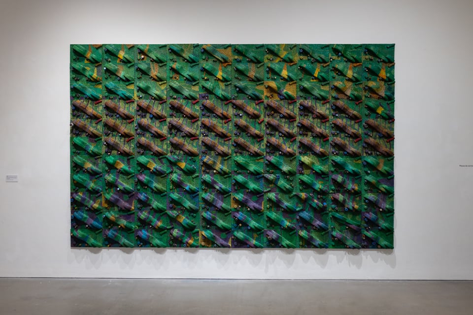 A photo from perhaps 10 feet away of a sculpture hanging on the wall. It is a green and blue and red and orange material with an undulating surface