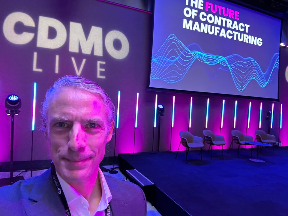 White man in gray suit & white shirt in front of stage for CDMO LIVE. The lighting is blue & purple