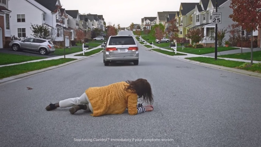 A woman in a yellow sweater is lying face down in the middle of a quiet residential street while a vehicle approaches in the background, about to run her over.