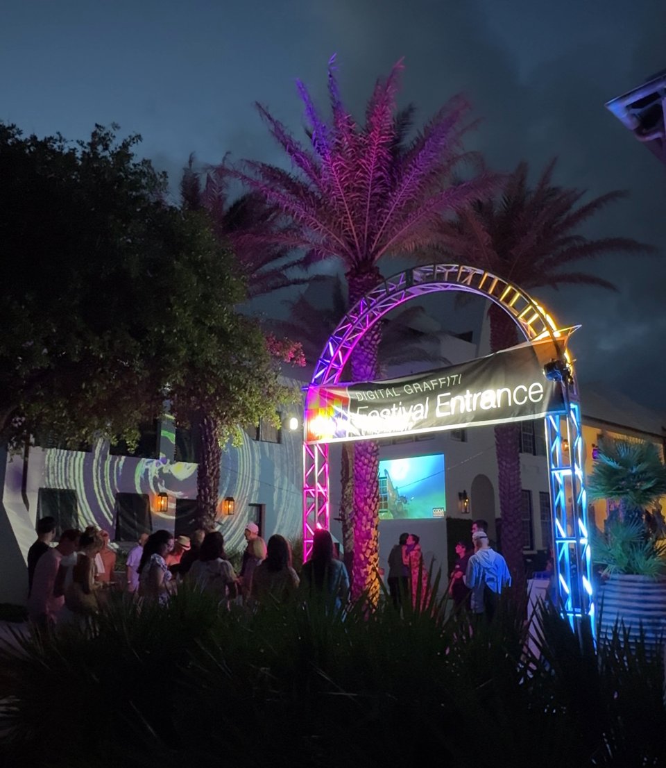 Night scene with palm trees and scaffolding entrance where a banner hangs that says Digital Graffiti Festival Entrance