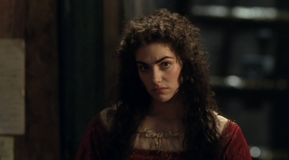 A pale young woman with curly, dark brown hair and thick, dramatic eyebrows. She is staring intently ahead, with an expression that is somewhere between a pout and a sneer