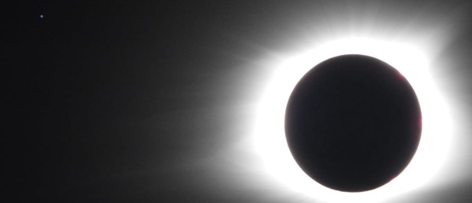 A photo of the 2017 eclipse, the dark disk of the moon surrounded by a bright white corona and some small points of red edging, and the faint blue dot of the SOHO spacecraft on the upper left