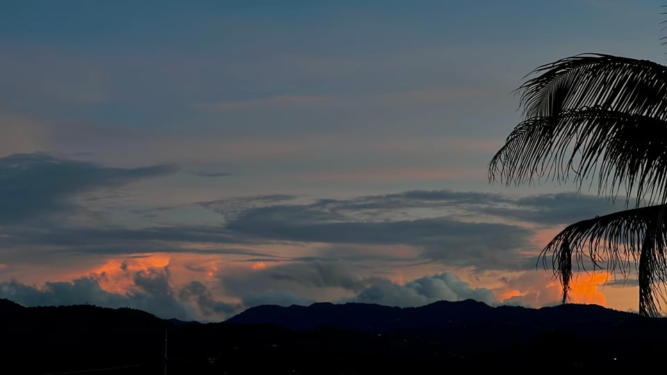 A sky and mountain landscape early in the morning. The sky is dark, the mountains and a palm tree are silhouetted, but the clouds in the very back are lit vibrantly
