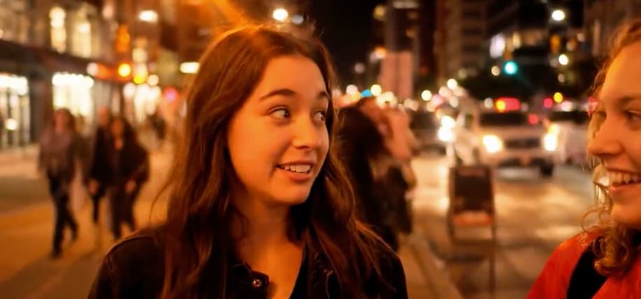 A young woman on a city street, with a smile and raised eyebrows, looks to her left at another young woman, seen in profile.