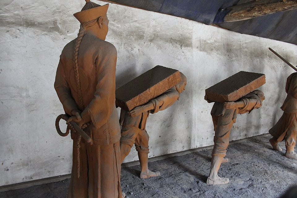 Three statues, one man with a long braided ponytail standing with his arms behind his back holding a whip, two smaller ones bent over with large stone slabs on their backs. They appear to be forced laborers.
