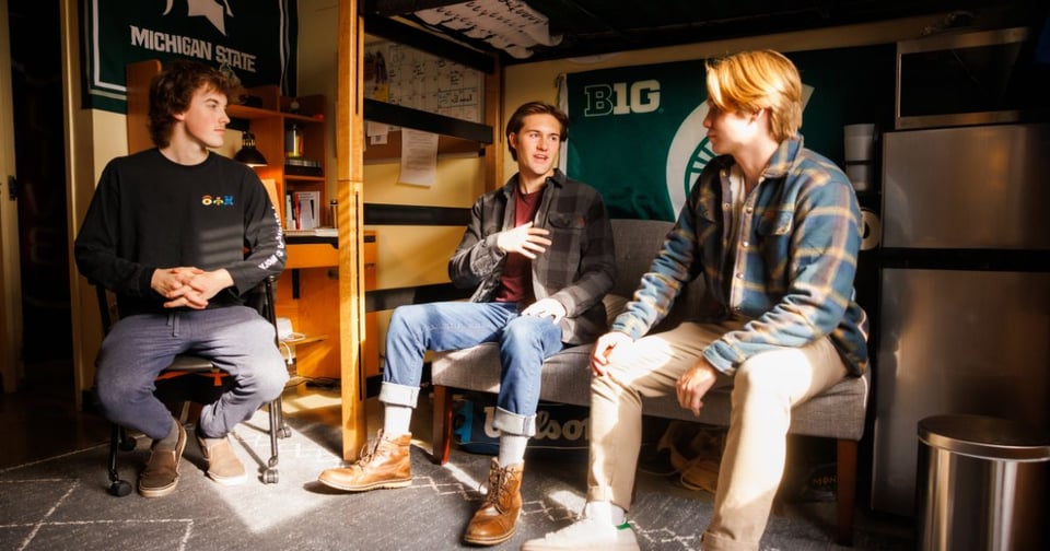 Three young men are seated in a college dorm room, engaged in conversation. The setting includes Michigan State University and Big Ten banners, indicating their school affiliation.
