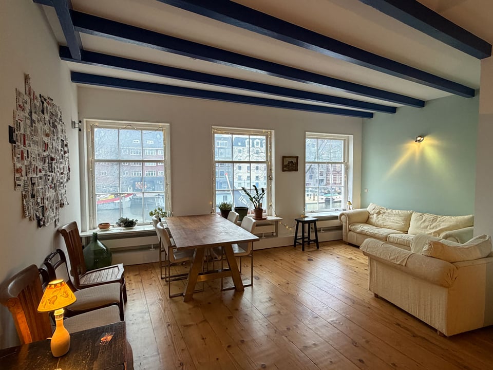 A dining room / living room with three large windows casting light onto the wooden floor.