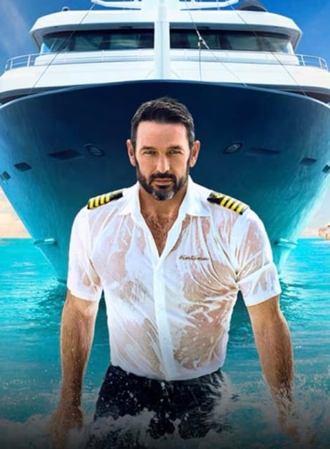 A person emerges from water wearing a captain's outfit; a boat is behind him