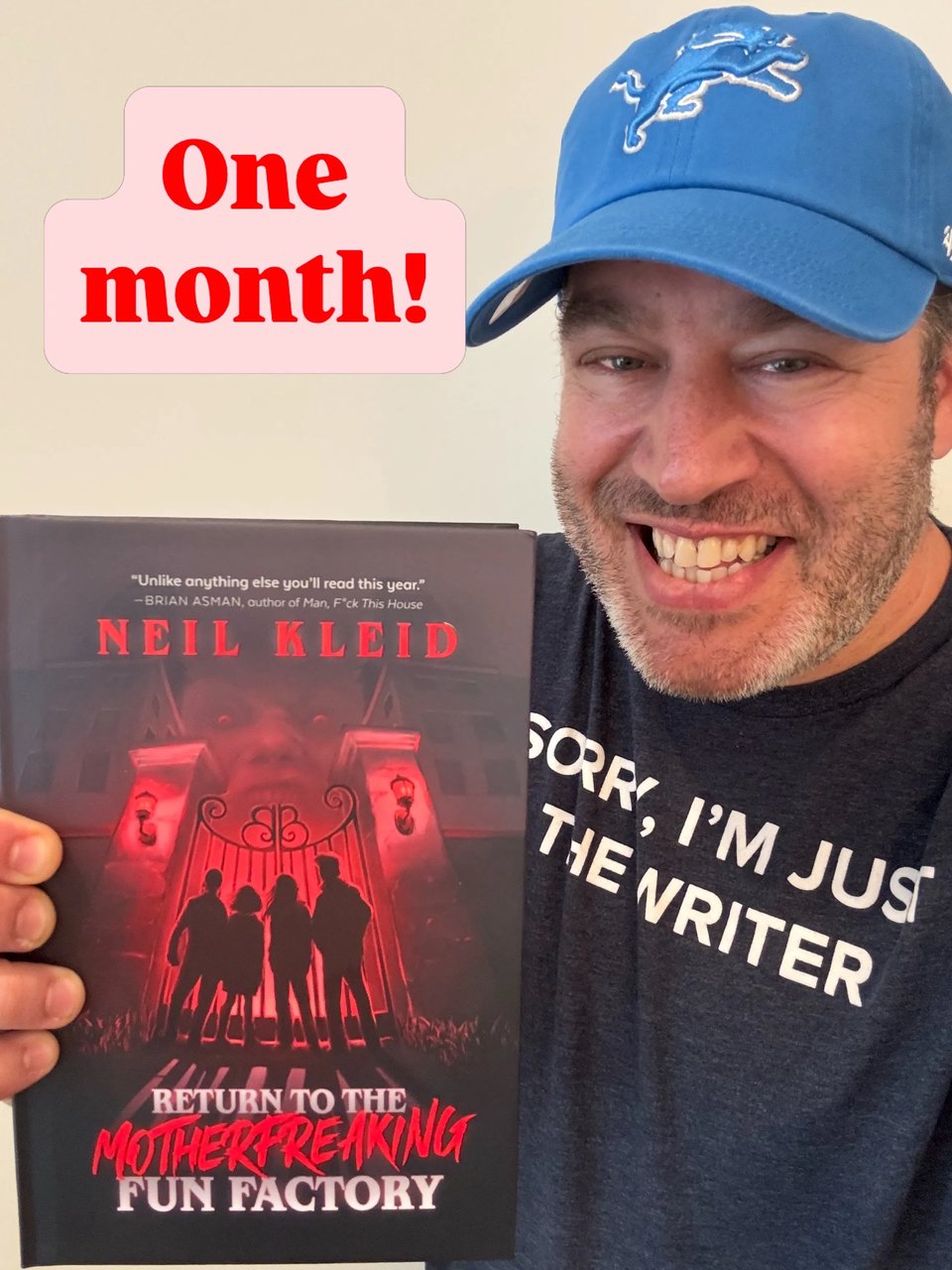 Neil in a blue Detroit lions baseball hat and a navy shirt with white text saying “sorry I’m just the writer” holding up a copy of his book, return to the motherfreaking fun factory” and red text that says “one month!"