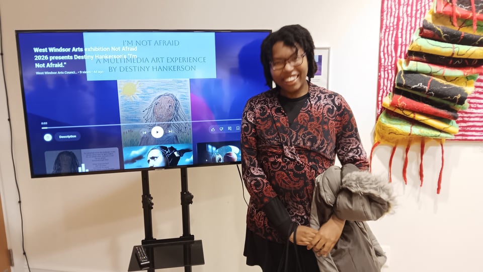 A photo of a young African-American woman at an art show, standing beside her digital art on the screen. She wears a patterned shirt and holds a coat, and her hair is in twists.