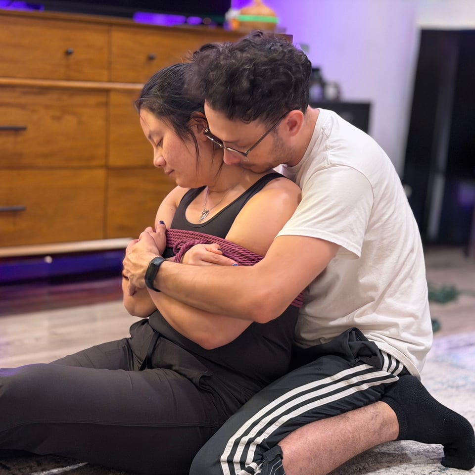 "A tender photo of two people sitting on a rug indoors. The person in front, wearing a black tank top and dark pants, sits with their eyes closed peacefully, their arms bound against their chest with purple rope. The person behind, wearing glasses, a white t-shirt, and track pants, kneels and wraps their arms tightly around the first person in a comforting embrace, resting their face against their neck.