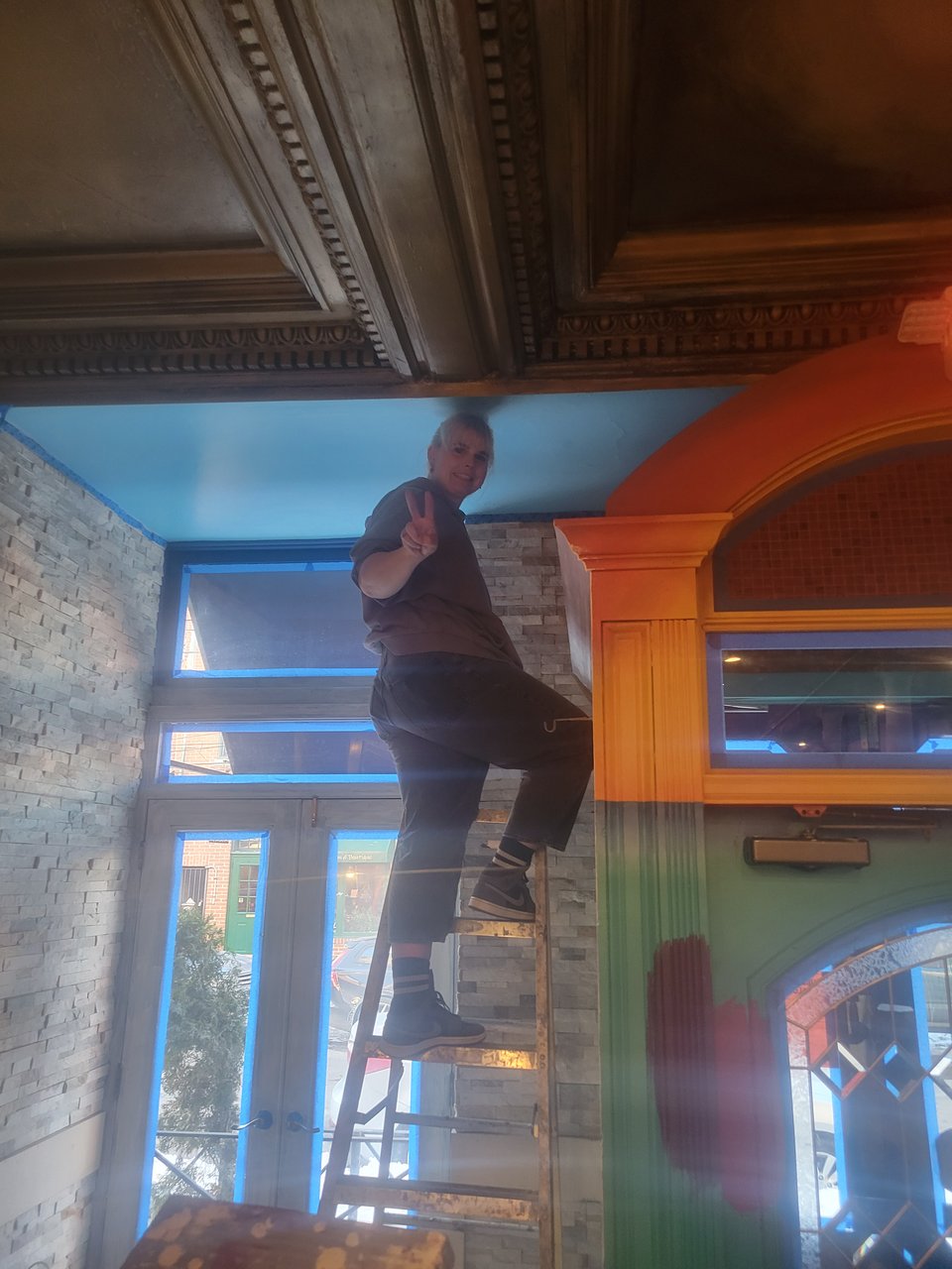 Someone on a ladder painting the ceiling, posing with a peace sign for the camera.