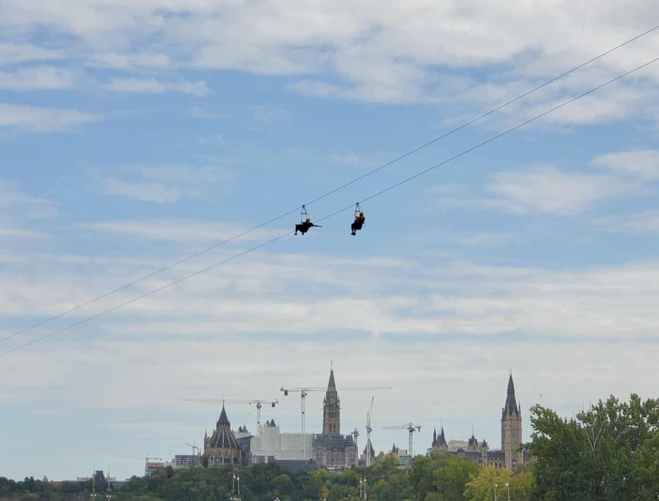 Two figures on a zipline framed above the Parliament Building in Ottawa, Canada. One has their arms spread wide. One clings to the harness above them.