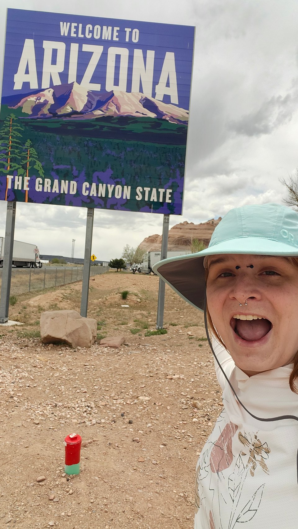 Arizona, a white woman in a blue hat, smiles and points at a sign that says "Welcome to Arizona: The Grand Canyon State"