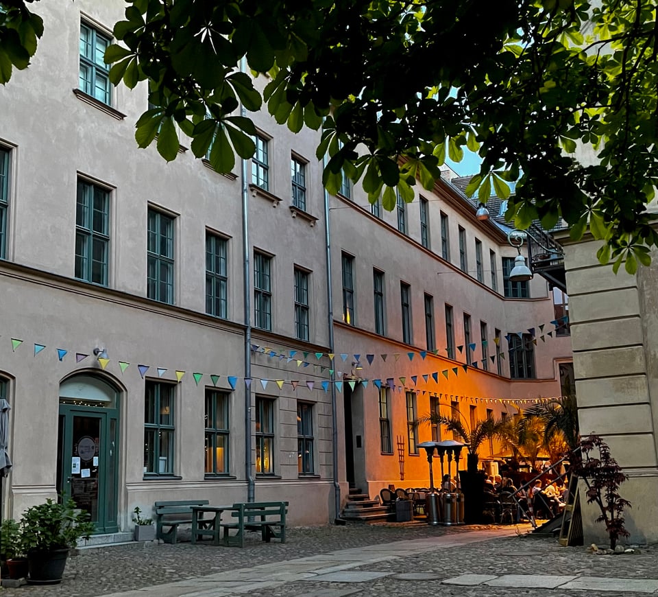 A residential building with a shop and a cafe/eatery on the ground floor. It is evening and the people visiting the cafe are eating with a glow of orange light near them. The whole picture is arched by branches of a tree at the top