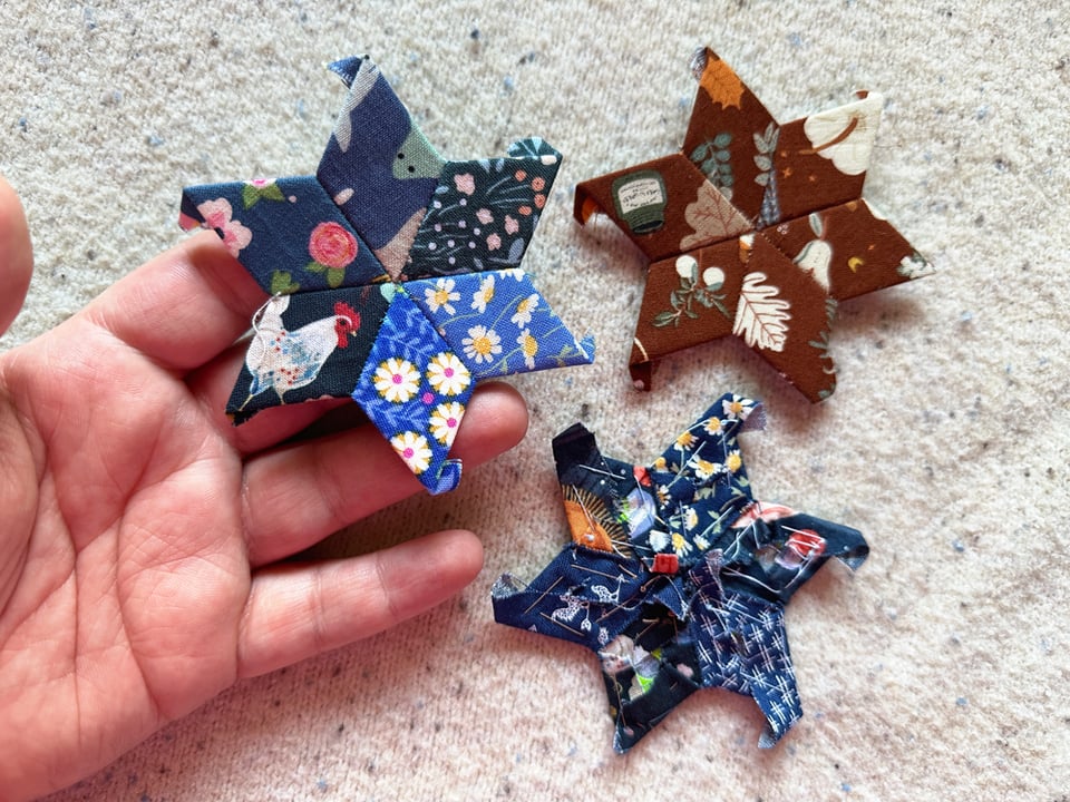 A trio of scrappy fabrics pieced together into star shapes. One cluster is brown, while the other two are in shades of blue. Jessi's hand holds up one of the blue ones.
