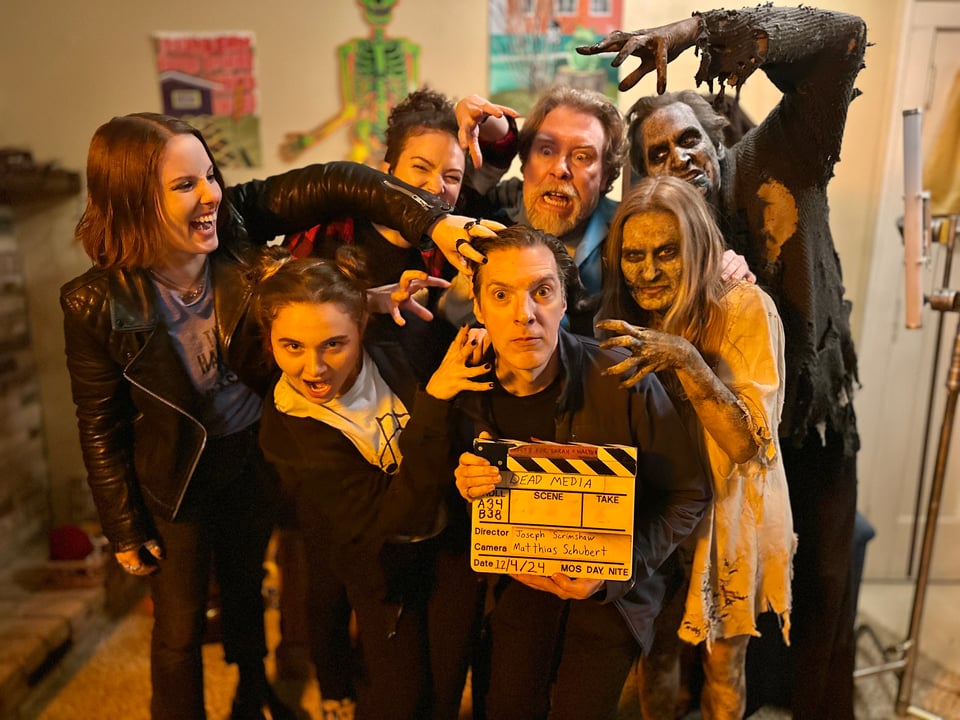 A small director with a slate that reads Dead Media, surrounded by cast members pretending to attack him