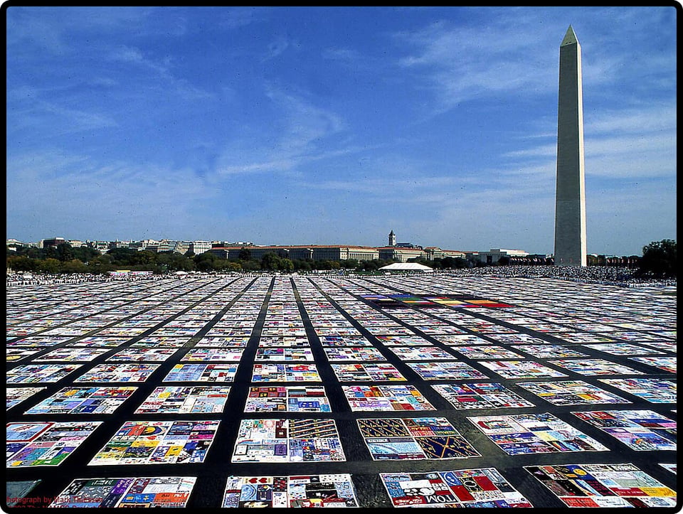 Photograph of the AIDS memorial quilt; the quilt squares stretch across the visible land in front of the Washington Monument