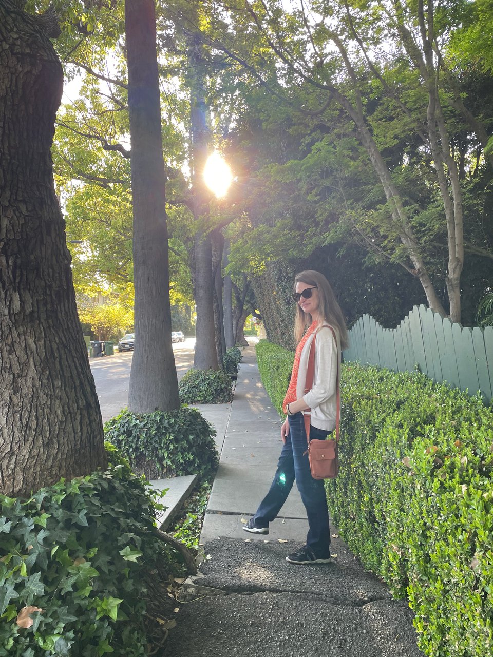 A woman pauses looking back toward the camera on a sunny, tree-lined residential street in Hollywood