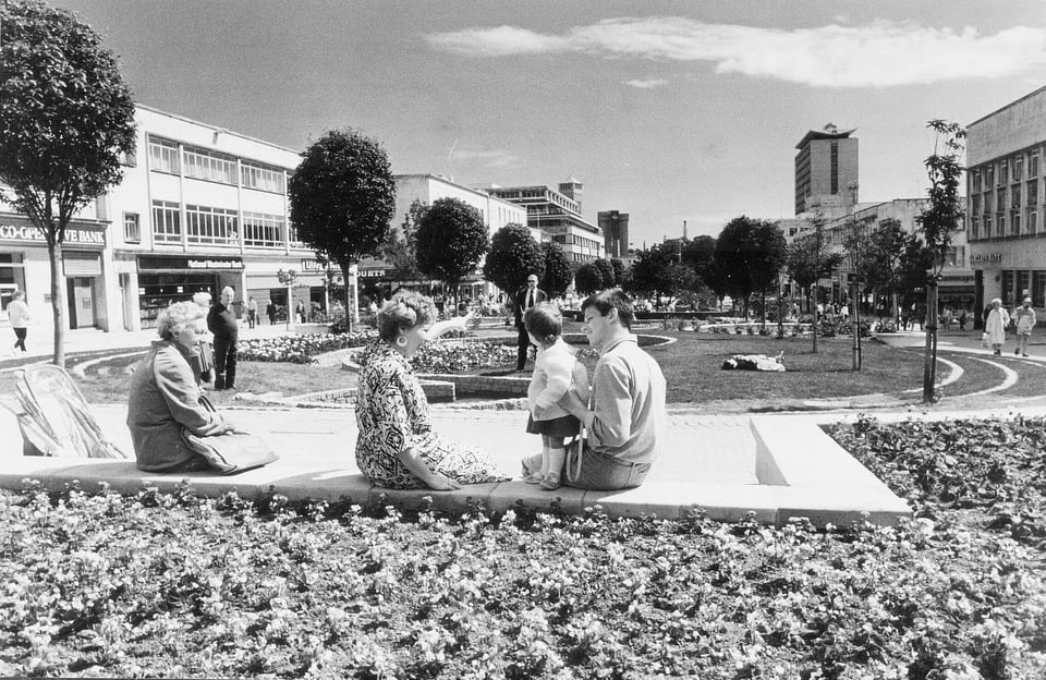 Black and white photo of Plymouth's Armada Way. A family is sat on the concrete edge of a huge flower bed. The woman is pointing a toddler's attention towards the Hoe in the distance. The whole avenue is wide, with three-storey buildings and young trees lining it.