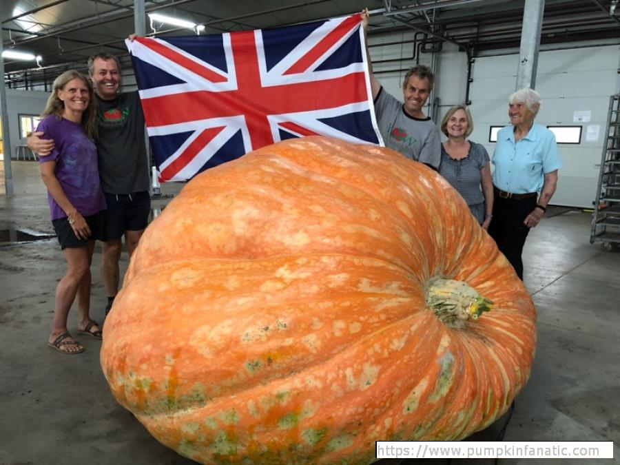 A photo of five people standing in an industrial room behind a gigantic pumpkin. The pumpkin looks to be about 5 feet In diameter, its massive, and slightly slumping downwards as if collapsing under its own weight. The people behind it are holding up a British flag and smiling