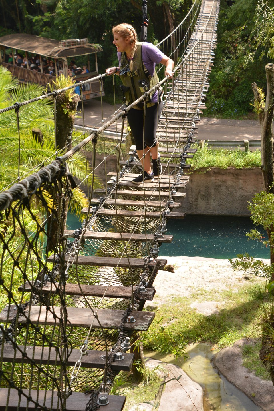 Cass, a 40 year old woman in black capri leggings and a purple t-shirt, strapped into a harness as she walks over a faux-rickety swinging bridge. She's looking off to her right, grinning broadly because she can see hippos.