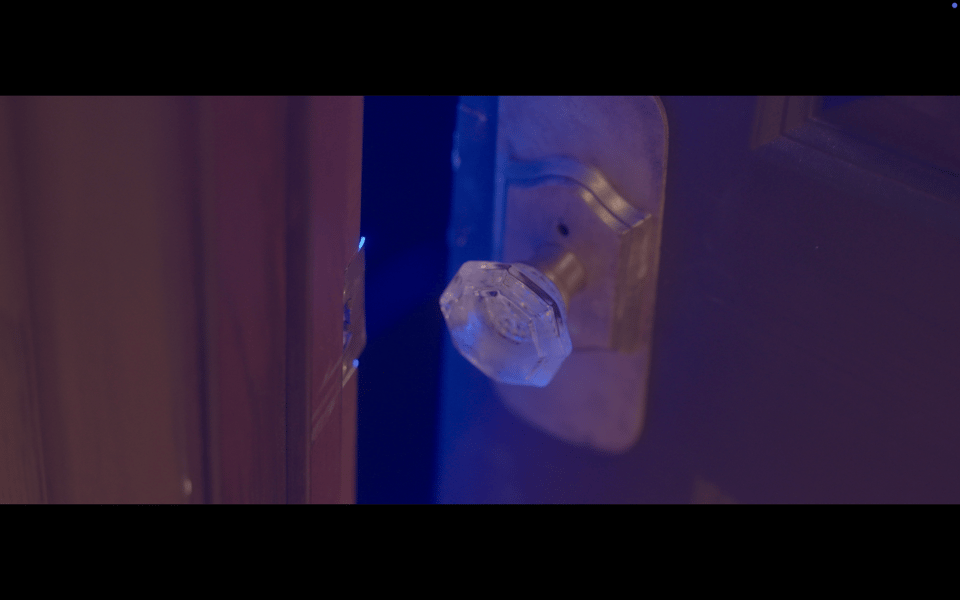 A frame of film showing an old door and knob creaking open, exposing eerie blue light
