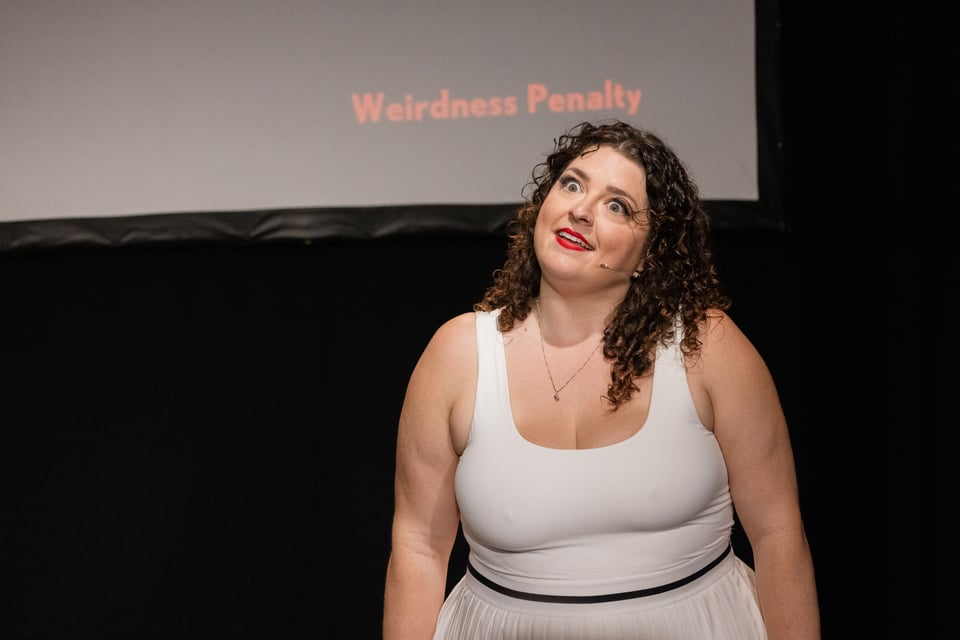 Photo of Mel making a funny grin in front of a projection screen. The screen reads "Weirdness Penalty" in red letters.