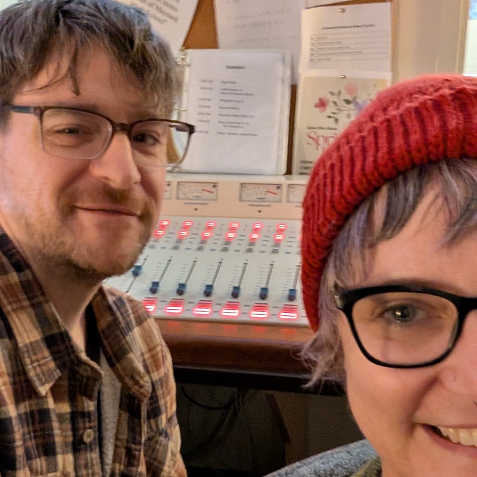 A selfie of a man and a woman in a radio booth. Both are wearing glasses and smiling in front of a sound board.