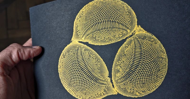 A pen plotted design with golden ink on white paper, held by a hand. Resembles three lemons connected forming a circle.