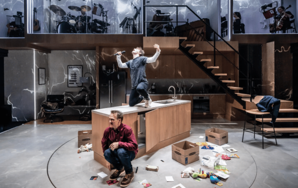 A performer dramatically poses with a microphone on a kitchen island while another figure sits on the floor amidst scattered boxes and debris, with a band visible on an upper level of the set.