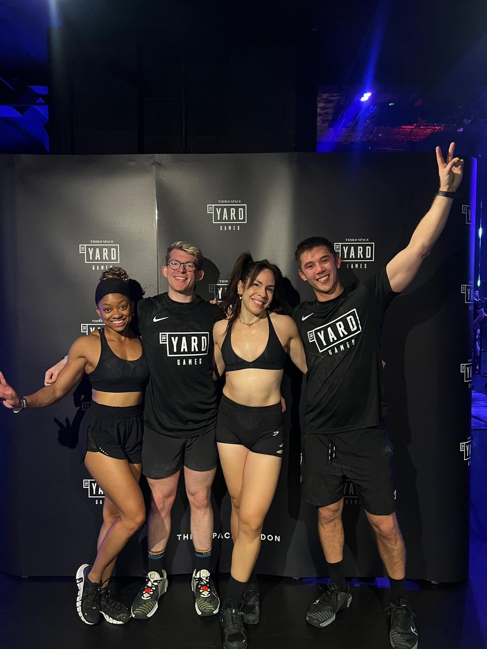 Four people wearing black athletic clothing pose together in front of a backdrop that reads “The Yard Games.” One person holds up a peace sign, and the group is smiling.