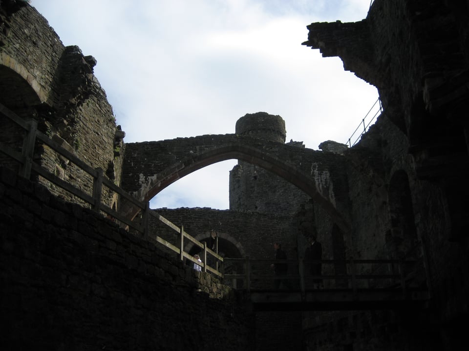 The shadowed interior of a ruined castle. Arches, one broken, rise overhead and a wood-railed passage is above.