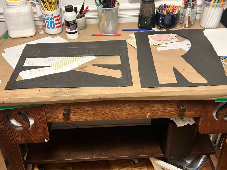 Large stencils of the letters R and K in warm grey green on an old wooden work bench.