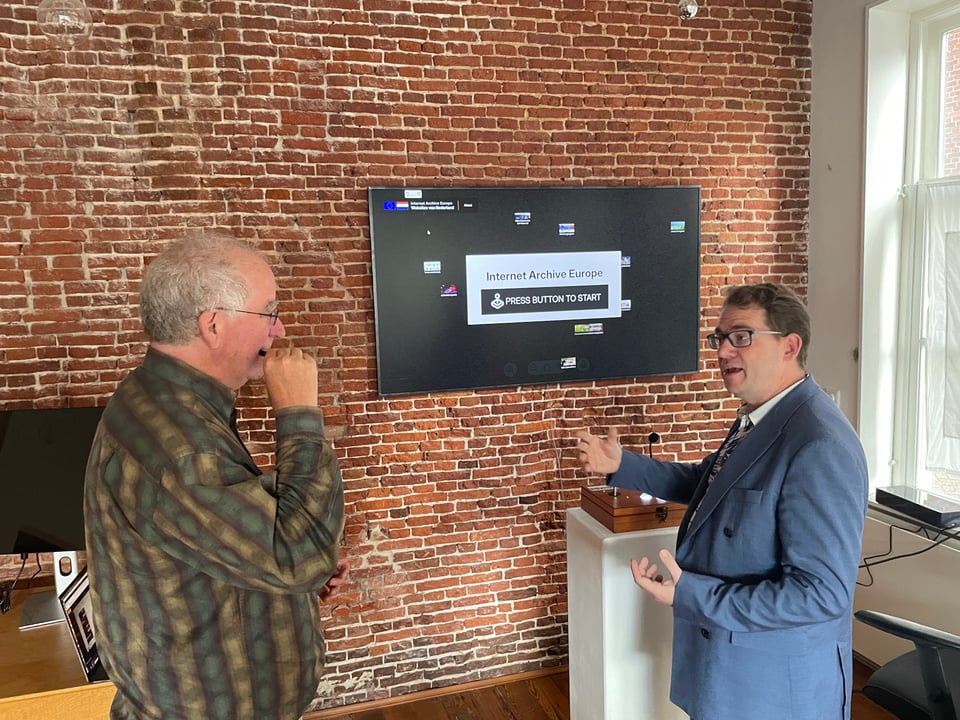 Two men discuss the finer points of web archiving in front of a large screen on a brick wall.