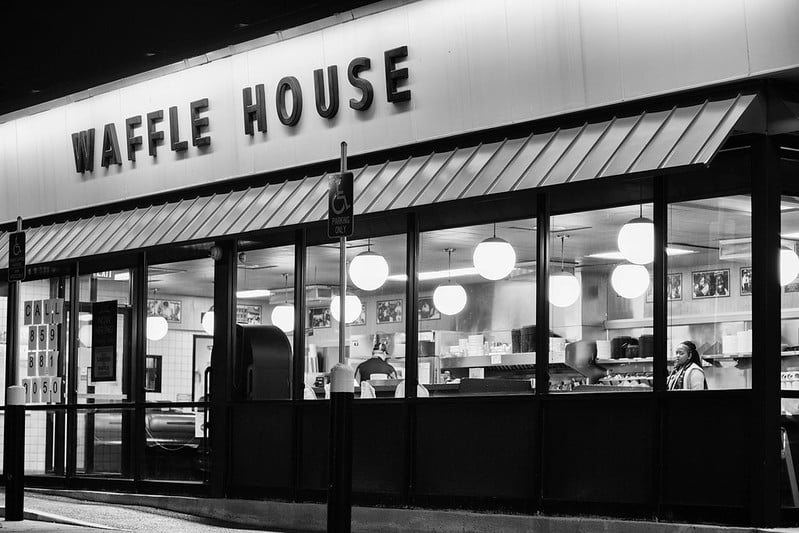 B&W photo shows the exterior of a Waffle House at night. The interior is empty except for the cook and one customer/