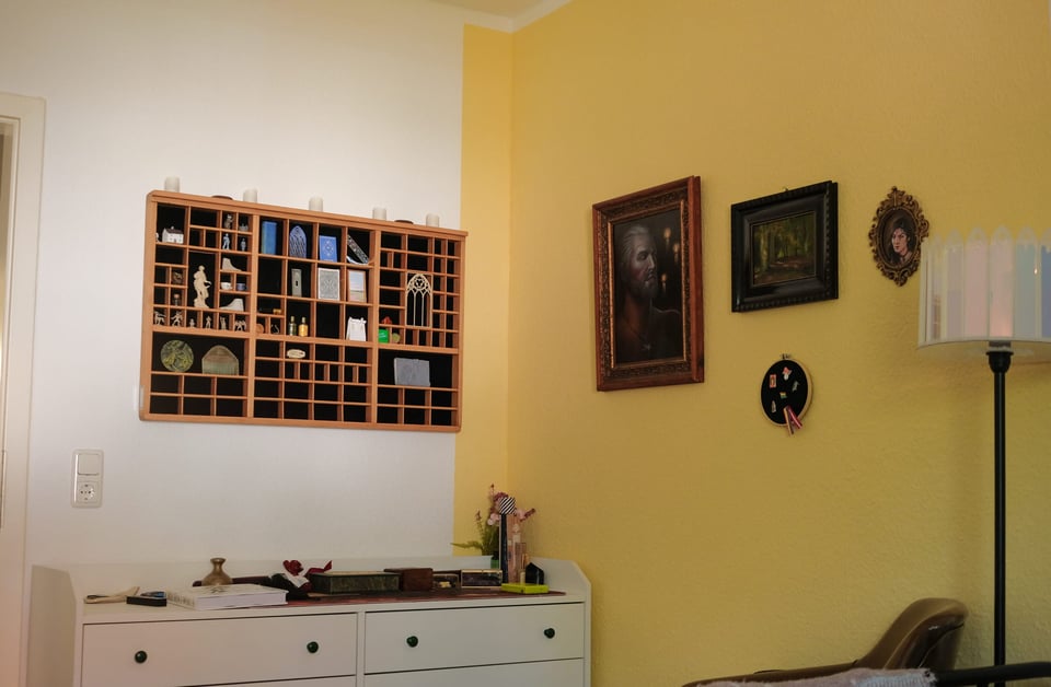 A photo of the corner of my bedroom. The trinket shelf hangs on the wall to the left, and three paintings and a pin frame on the wall on the right. The right wall is a pale yellow, and the left wall is still white.