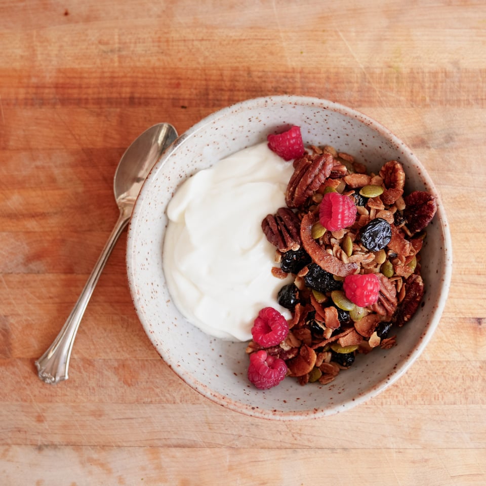 A bowl of yogurt with granola and fruit on the top sits on a wooden table with a spoon.