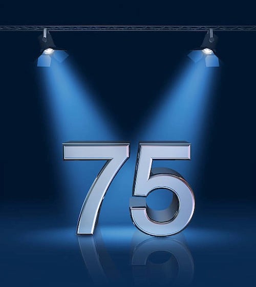 The number 7 and the number 5, tall shiny and metallic, on a smooth glassy blue stage. Each is lit, spotlight-style, by a separate fresnel stage light hanging from above.
