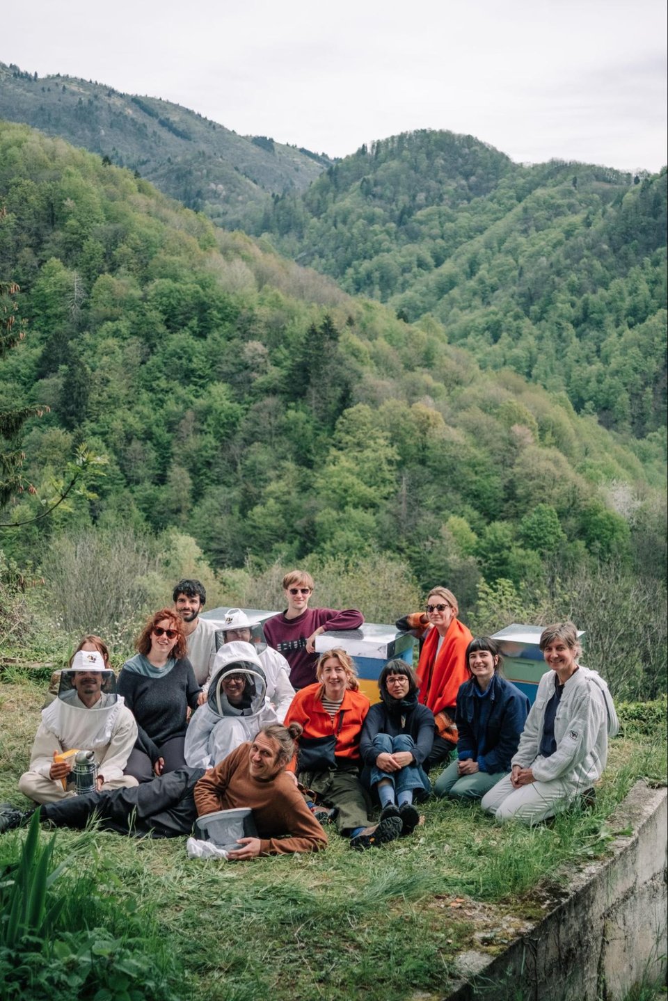 [A group of 12 people, three wearing bee-keeping outfits, surrounded by mountains covered in lush trees.]