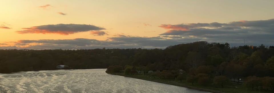 A photo of sunrise over a rippled bend in a river, lined with trees. Above and beyond the river float a collection of orangey-pink and slate blue puffy clouds. The sky behind the clouds runs from a pale yellow on the left side and gradates into a greenish gray.