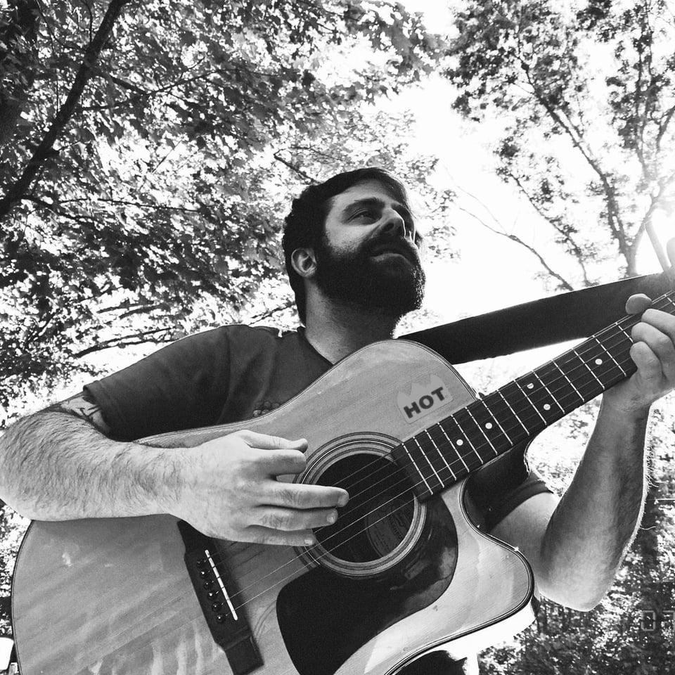 Looking up at a bearded man playing an acoustic guitar with a sticker that says "hot" on the body just above the neck.