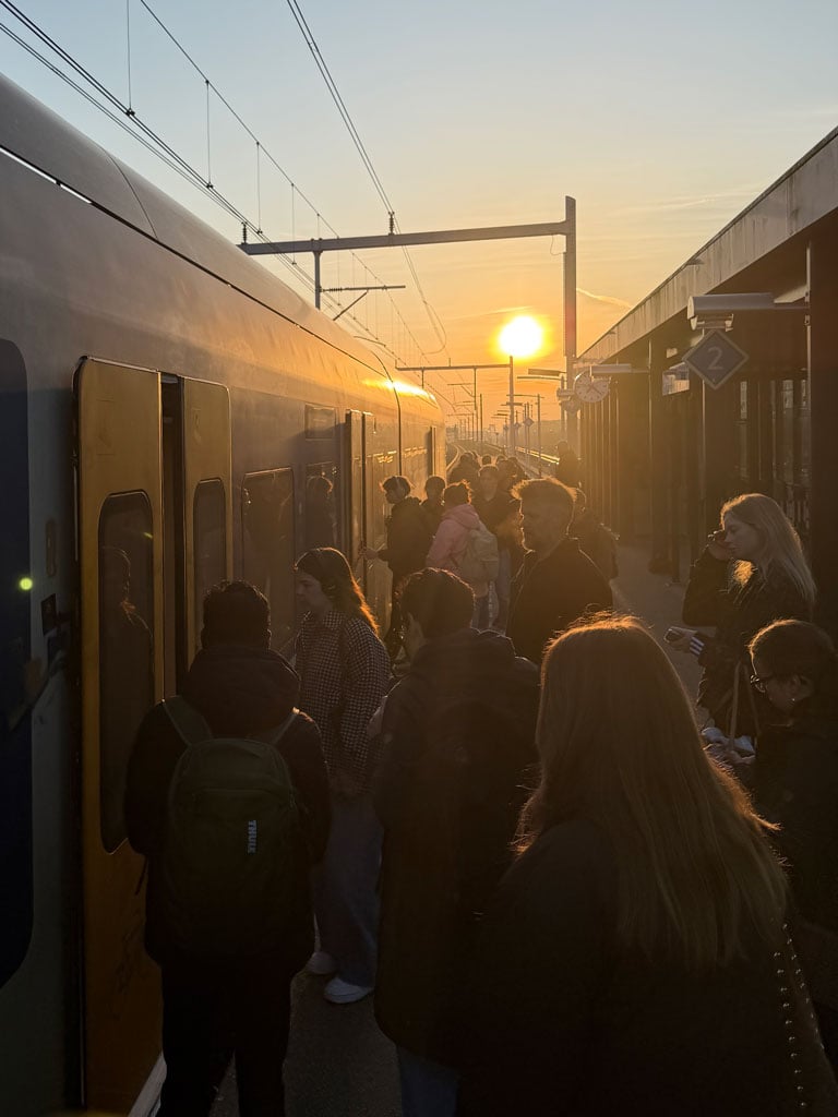 Many commuters gather in front of the train to board in the early morning as the sunrise shines on the horizon.