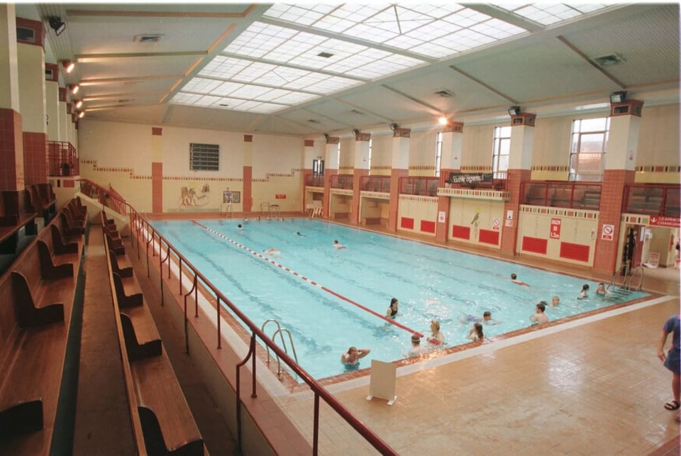 A very 1980s colour graded photo of the inside of the building. The 100 foot long pool is still under the 100 foot long glass roof, and the ranked spectator seating is still well out of splashing range. At the far end, the diving platforms have gone. In place there is a large mural on the tiles with what looks like the Egyptian god Ra. Who I was not aware was a big swimmer.