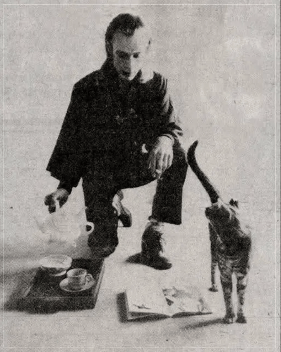 A black and white photo of Brian Eno kneeling on one knee and pouring a cup of tea with his cat in front of him looking back. The photo is titled: Brian Eno pours tea for his cat, 1974