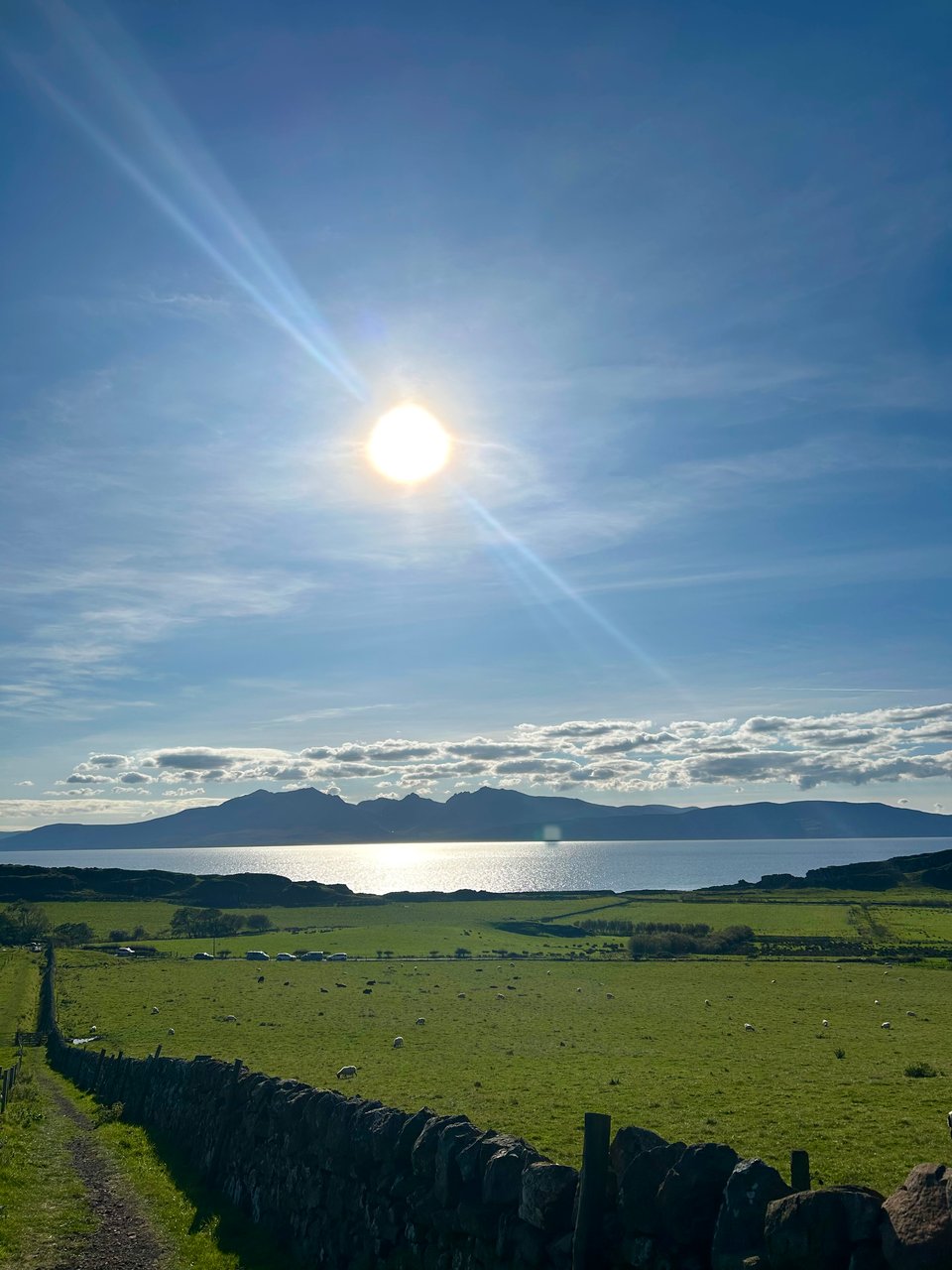 The Isle of Arran is looking very craggy in the distance, across a glittering sea. There are green field in the foreground, dotted with sheep. There's an ancient stone wall running from the far right close up to the far left in the distance. Image by Rowan Ambrose.