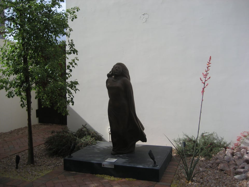 A statue of a woman in a flowing dress, head tilted to look up at the sky, carved from black stone and on a black plinth in front of a white wall.