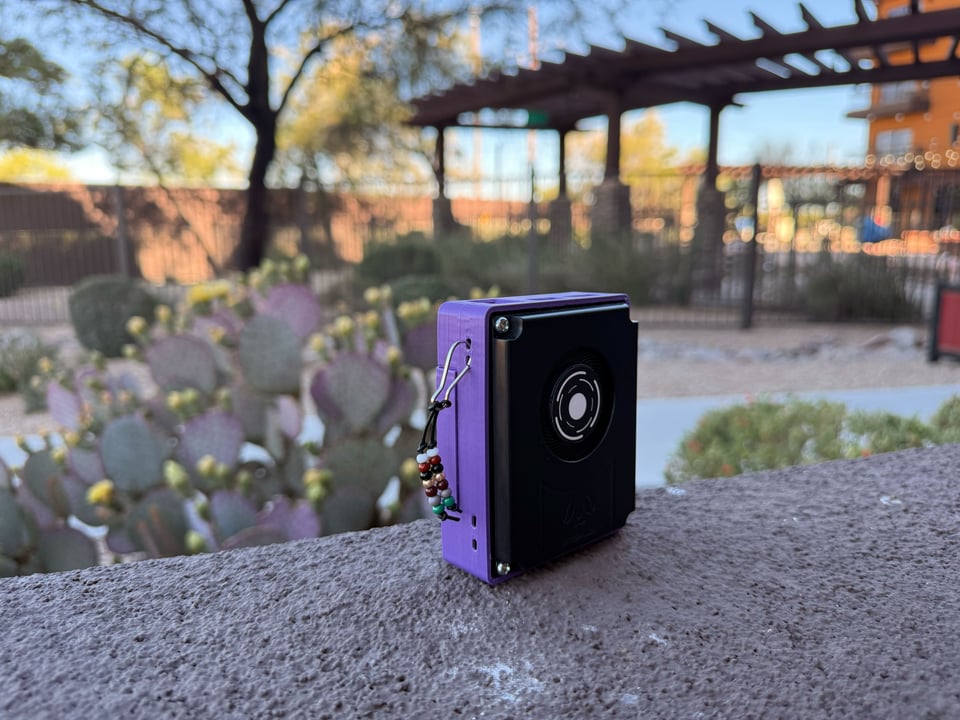 A small purple and black box with beaded tassels on the side (a steady state hard drive thats running Niiwin) sits on a stone wall overlooking a patio structure with cactus in view.