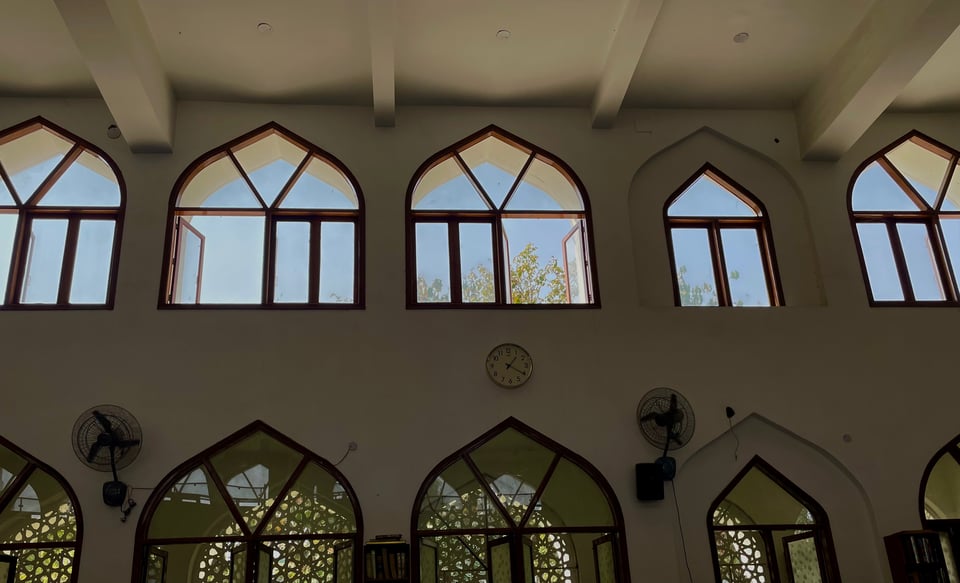 A shot of high ceiling and huge windows of a mosque, on a 30 degrees afternoon in delhi. The bottom row of windows have an intricate Arabesque geometric breeze block wall. The top row of windows show the tops of trees and blue sky beyond them