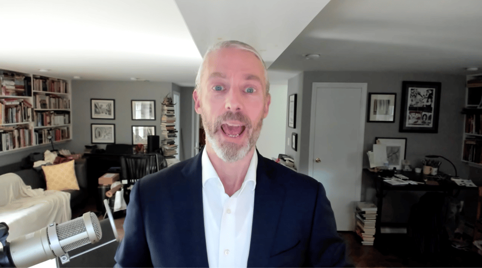 photo of a white man in a blue suit and white shirt, speaking to a webcam. His mouth is open.